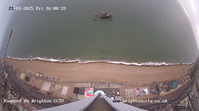 Aerial view of a sandy beach with rolling waves and a cloudy sky. In the distance, a dilapidated pier is visible standing on the water. The shoreline is dotted with various structures, including colorful beach huts and amusement rides. The scene is calm, with a few people walking along the beach near the water's edge. The date and time are displayed at the top of the image.