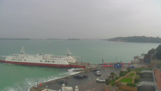 A view of a harbor with a large white and red ferry docked near the shore. The water is a greenish-blue color and has some waves. In the background, there are several small islands visible on the horizon, under a gray sky. In the foreground, there is a parking area with a few cars and some greenery, along with a blue structure and red barriers.