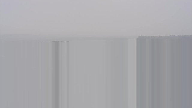 A hazy landscape with a low visibility scene, featuring soft gradients of grey and white. The horizon is faintly visible, blending into a cloudy sky. There are indistinct shapes of distant landforms or trees along the water's edge, with no distinct outlines due to the fog. The overall atmosphere appears calm and muted.
