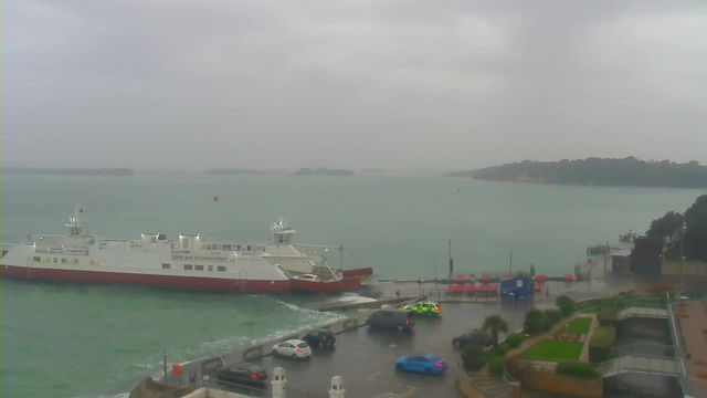 A foggy waterfront scene shows a large white ferry with a red bottom docked at a terminal. The water is choppy with waves, and distant green hills are visible across the bay under a gray sky. Several cars are parked near the terminal, including a blue car and emergency vehicles. The area appears wet, indicating recent rain. On the dock, there are red objects and a small blue building in the background.