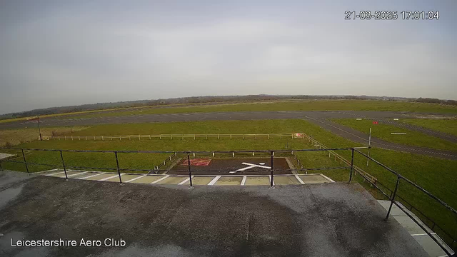 A view from a webcam showing a grassy airfield with a paved runway in the background. In the foreground, there's a railing that appears to be part of a viewing deck. The sky is mostly cloudy, and a few trees can be seen in the distance. A red flag is placed near the edge of the runway, and there is a large white marking on the ground resembling a cross. The scene has an overall calm and open atmosphere.