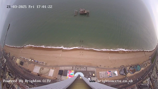 Aerial view from a high vantage point overlooking a beach. The sandy shoreline meets the gently rolling waves of the ocean. In the distance, a partially submerged pier is visible. Below, there are sections of a boardwalk with colorful geometric patterns and scattered activities. The sky is overcast, creating a muted light over the scene. The date and time are displayed in the upper left corner, with a watermark for Brighton CCTV in the bottom left.