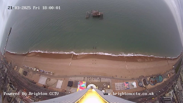 A wide-angle view from a high point overlooking a beach. The image shows a sandy shoreline with gentle waves lapping at the edge of the water. A wooden pier is visible in the water, partially submerged. The beach is lined with various structures, including amusement rides, colorful patterns on the ground, and what appears to be a swimming pool. The sky is mostly cloudy, and the scene captures a sense of a coastal environment at dusk. The date and time are displayed at the top left corner of the image.