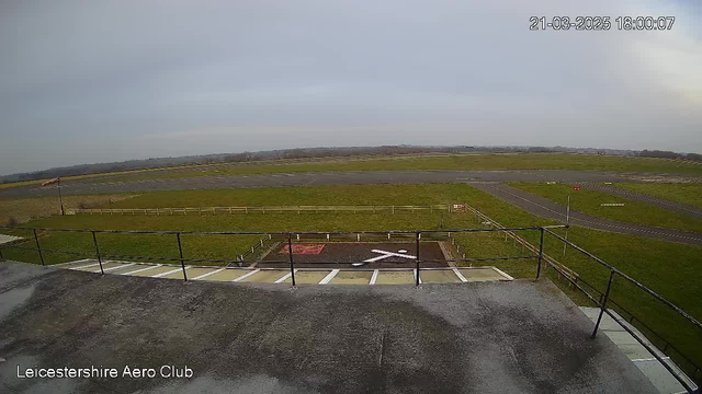 A view from a webcam at Leicestershire Aero Club showing a grassy airfield with a runway in the background. There is a circular helipad marked with a white "X" in the foreground. A fence lines the edge of the field, and a windsock is visible to the left, indicating wind direction. The sky is overcast with a soft grayish hue, and the scene is devoid of any aircraft or people.