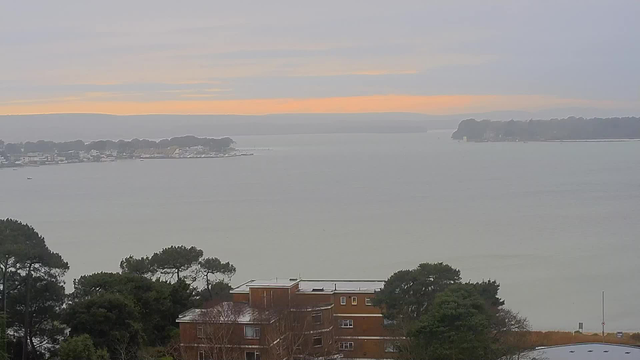 A hazy view of a calm body of water, with distant land on either side. The sky is overcast with soft gray clouds, hinting at an orange glow from the setting sun near the horizon. In the foreground, there is a brown multi-story building partially obscured by trees, and some boats are visible in the water. The atmosphere appears tranquil and slightly moody.