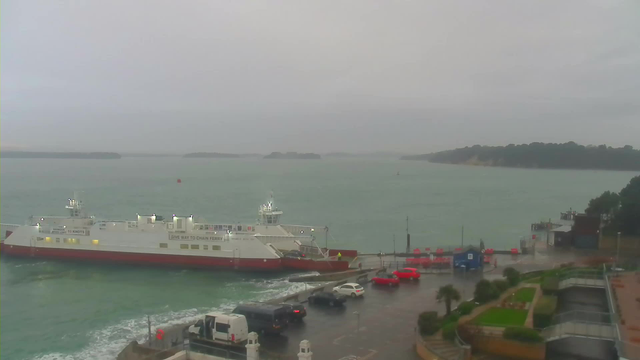 A ferry is docked at a waterfront terminal on a cloudy day. The water is choppy, reflecting gray skies, with several small islands visible in the distance. In the foreground, there are parked vehicles, including red cars, near the terminal. The area has a few trees and landscaped sections, indicating a developed shoreline. The atmosphere appears calm despite the overcast weather.