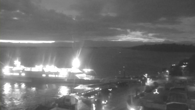A nighttime view of a waterfront scene. A large ferry boat is docked, glowing brightly with lights against the dark water. The water reflects the lights, creating shimmering patterns. In the background, clouds are visible, and the sky is partially illuminated by a horizon light. The shore is lined with buildings and boats, and there are some parked vehicles in the foreground.