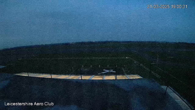 A dimly lit scene captures a wide view of an airfield at dusk. In the foreground, there is a flat rooftop with a light-colored edge, possibly indicating a structure below. The field is mostly dark with a few white runway markers visible. The sky is a gradient of deep blue transitioning to lighter shades near the horizon. The timestamp indicates the view was captured on March 21, 2025, at 19:00:31, and a label reads "Leicestershire Aero Club" in the bottom left corner.