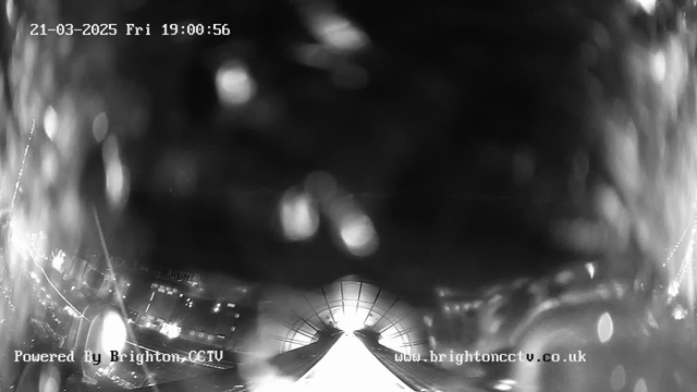 A blurred black and white image taken from a webcam, showing an unclear scene with bright reflections and light spots. The lower portion of the image features a pronounced white area, suggesting the presence of a pathway or road. At the top, there are faint outlines that hint at buildings or structures in the background. The timestamp in the upper left corner indicates the date and time as March 21, 2025, at 19:00:56. A watermark with "Powered By Brighton CCTV" is visible in the bottom left, along with a website URL in the bottom right. The overall clarity is poor, making specific details indistinguishable.