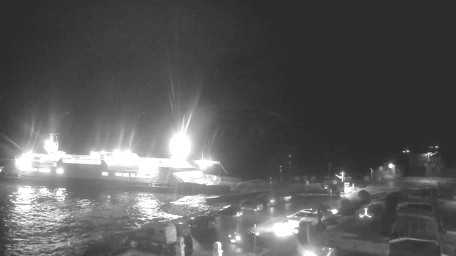 A nighttime scene showing a large illuminated ferry docked at a pier. The ferry is brightly lit with multiple lights, creating reflections on the water surface. In the foreground, various vehicles are visible near the pier, illuminated by their headlights. The surroundings are dark, indicating it is nighttime. The water appears to be calm.