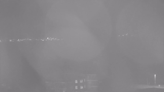 A low-contrast black and white image showing a foggy scene. In the background, there are faint lights scattered along a horizon, possibly representing distant buildings or streetlights. The foreground appears dark, with indistinct shapes that could represent structures. The overall visibility is limited due to the fog, creating a muted and hazy atmosphere.