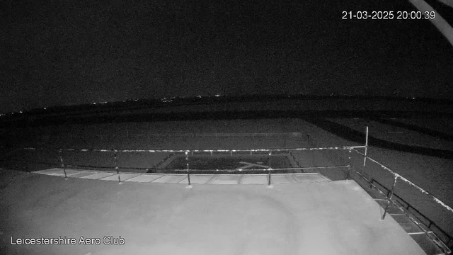 A dark nighttime view from a webcam at the Leicestershire Aero Club. The image shows a flat rooftop with a railing in the foreground, and a runway in the background. There are faint lights visible in the distance, likely from buildings or vehicles. The overall scene has low visibility and is predominantly dark. The timestamp in the corner indicates the image was captured on March 21, 2025, at 8:00 PM.