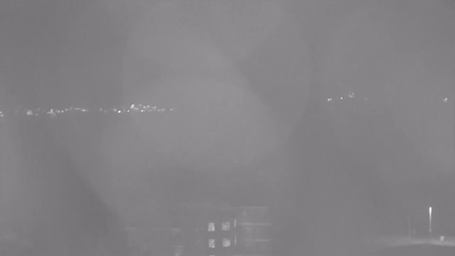 A blurred monochrome image from a webcam showing faint lights in the distance, likely representing buildings or streetlights. The foreground is largely obscured, creating an unclear view of the entire scene.