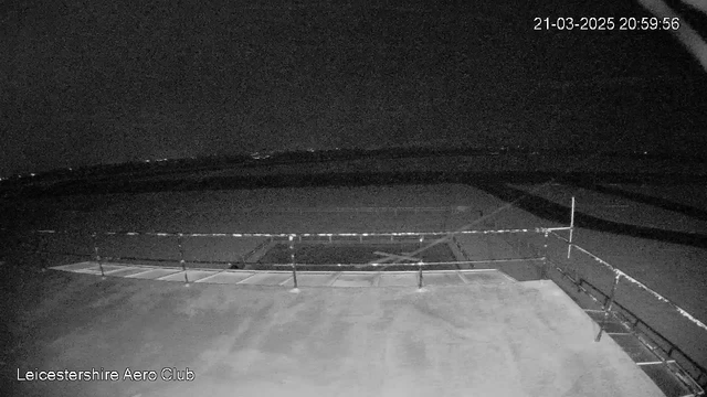 A dark scene captured from a webcam at Leicestershire Aero Club. The foreground features a flat, empty surface with a railing. In the background, faint lights can be seen in the distance, indicating a nighttime landscape. The overall image is predominantly black with minimal visibility. The date and time are displayed at the top right corner.