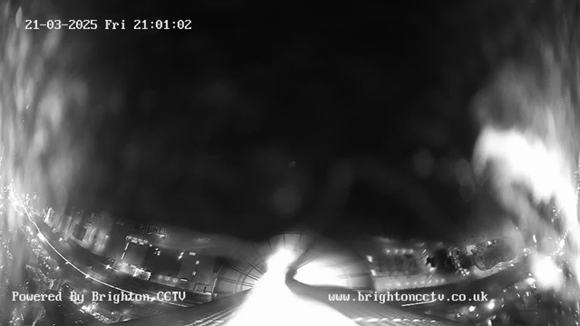 A black and white webcam image showing a dimly lit urban landscape at night. The foreground includes a street with some visible lights and buildings, while the background is mostly dark with blurred elements. The date and time are displayed in the top left corner, reading "21-03-2025 Fri 21:01:02," and the bottom left corner includes the text "Powered By Brighton CCTV," along with a website link. The overall scene appears hazy, suggesting potential rain or fog.