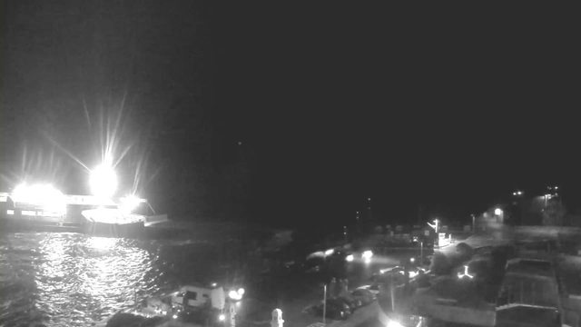 A dark scene with a bright light source on the left, likely from a ship or boat, reflecting off the water. The water appears choppy, and there are dim lights from small boats and structures on the shore. The background is mostly dark, indicating it is nighttime.