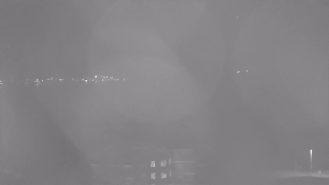 A blurred, low-contrast black-and-white image shows faint outlines of lights in the distance against a foggy backdrop. There are streaks that may represent reflections or raindrops on the lens. A building with a few illuminated windows is visible at the bottom of the image, contributing to the overall dim atmosphere.