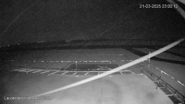 Black and white image of a dark landscape taken from a webcam. In the foreground, there is a flat surface with a painted symbol resembling a compass. A barrier or fence is visible along the bottom edge, dividing the image. The background shows a vast, empty area with faint outlines of distant hills or structures. The time and date are displayed in the top right corner, indicating it is nighttime.