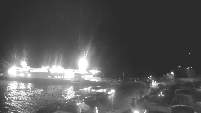 A nighttime scene of a harbor with a large, brightly lit ferry docked at the side. The water reflects some of the bright lights from the ferry. Several smaller boats are moored nearby, and the area is dimly lit with distant lights visible along the harbor. The scene has a dark, shadowy background, indicating a nighttime setting.