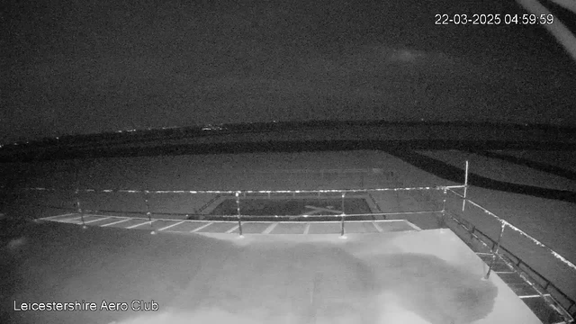A dark and shadowy scene captured from a webcam at Leicestershire Aero Club. The image shows a flat rooftop surface with a railing on the left side. In the background, there is a vast expanse of dark sky, possibly with faint lights indicating distant buildings. The date and time in the upper right corner show March 22, 2025, at 4:59 AM. The overall lighting is low, contributing to the dim ambiance of the nighttime view.