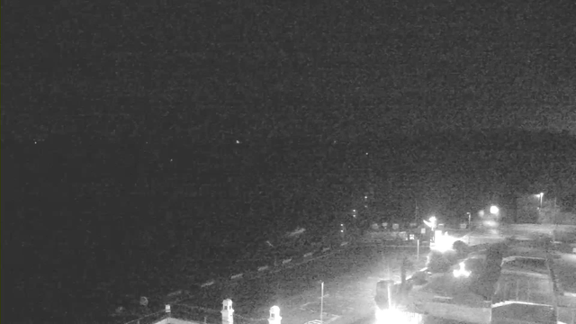 A dark scene captured by a webcam, showing a vague outline of a waterfront area. The horizon is mostly indistinct, with a few dim lights appearing sporadically. In the foreground, there are structures, possibly buildings or signage, illuminated by artificial light, while the overall atmosphere is shadowy and nighttime.