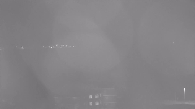 A dimly lit view from a webcam shows a foggy or misty scene. Some faint lights can be seen in the distance, indicating possible buildings or streetlights. The foreground appears dark with vague shapes, possibly representing structures, while the background is hazy and unclear.