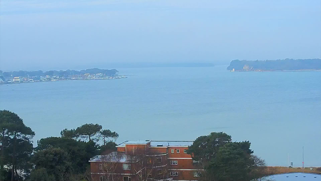 A serene landscape view featuring a calm body of water with light blue tones. In the foreground, there are several green trees. Below, a brick building with multiple windows can be seen. In the distance, a shoreline with boats docked is visible, along with a lightly foggy horizon. The sky is overcast, blending soft hues of blue and grey.
