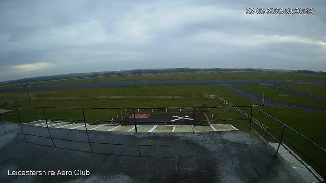 A view from the Leicestershire Aero Club webcam showing an expansive green field and an asphalt runway under a cloudy sky. In the foreground, there is a railing and a section of the deck. The grass area is divided by a small wooden fence, and there are markings on the runway. The overall scene is calm, with no visible aircraft.