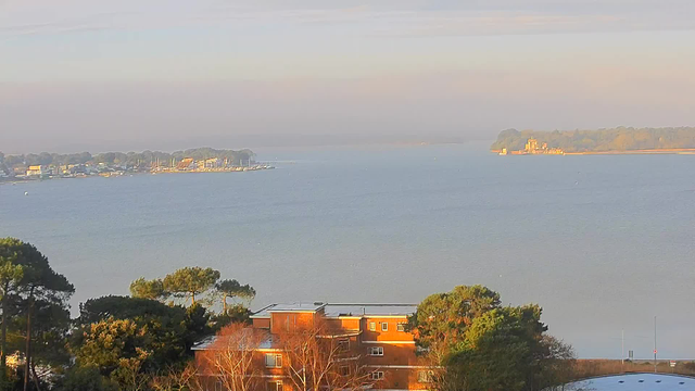 A view of a calm body of water under a clear sky, with soft morning light. In the foreground, there is a low, reddish brick building partially obscured by trees. To the left, several small boats are moored along a colorful waterfront lined with houses and businesses. In the distance, an island with greenery is visible, featuring a prominent structure that resembles a castle. The horizon is slightly hazy, adding a gentle gradient to the backdrop.