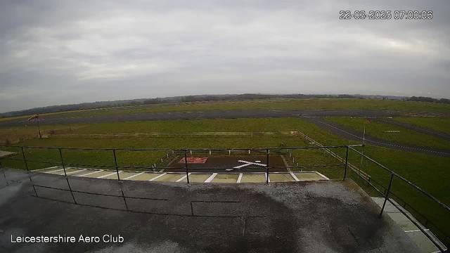 A view from a webcam at Leicestershire Aero Club. The foreground shows a tarmac area with a white 'X' marking on the ground, bordered by a railing. Behind it, a grassy field stretches out, with a paved runway visible further back. The sky is overcast with clouds, contributing to a gloomy ambiance. The scene is quiet and still, suggesting early morning conditions.