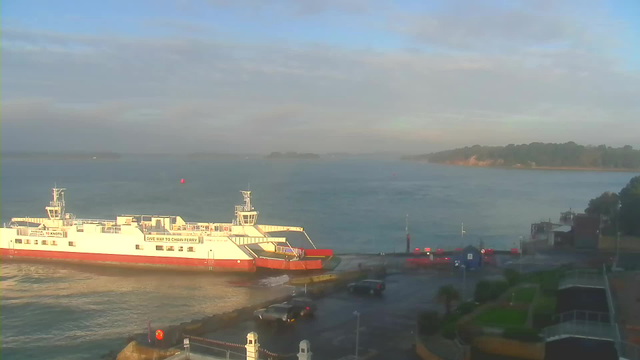 A ferry is docked at a harbor with a red and white color scheme, positioned near the water's edge. The background features a calm sea under a partly cloudy sky, with a few small islands visible in the distance. In the foreground, there is a paved area with parked cars and some greenery, including bushes and trees. A red buoy can be seen floating in the water nearby. The scene is well-lit, indicating it is either morning or late afternoon.