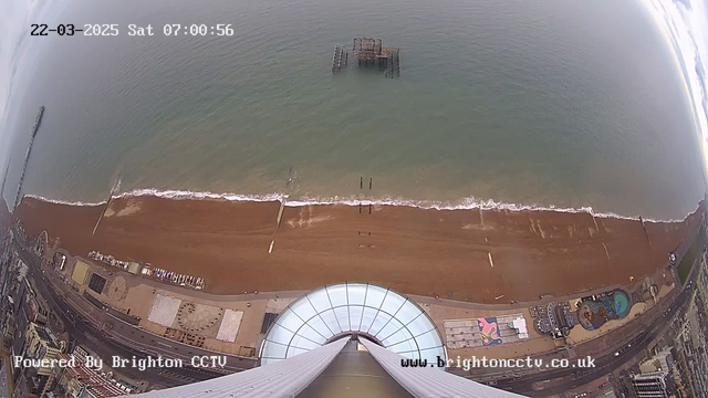 Aerial view of a beach with a wide expanse of sandy shore and gentle ocean waves. The scene shows a pier extending into the water on the left, while a partially submerged old pier structure can be seen in the distance. Along the beach, there are various structures, including amusement rides and a swimming pool area. The sky is overcast, and the time stamp indicates it is early morning.