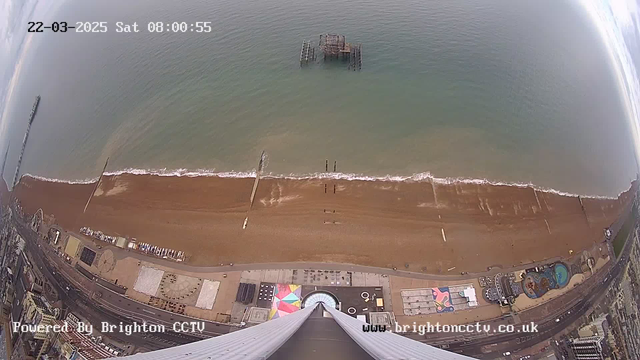 Aerial view of a beach with a sandy shore and gentle waves lapping at the edge. In the distance, there is an old pier structure extending into the water. The shoreline is lined with a promenade featuring various buildings and attractions, including a colorful circular design on the ground. The sea is calm, reflecting clouds in the overcast sky above.