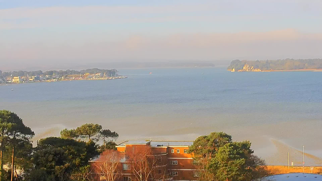 A scenic view of a calm water body under a clear sky, with low-lying clouds in the distance. In the foreground, there are green trees and a building with red brick. To the left, a shoreline is visible with various boats docked and moving. In the background, an island can be seen with some structures, likely a castle or old buildings. The water is a mix of blue and reflections of soft colors from the sky.