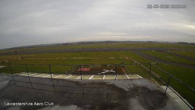 A view from a webcam showing a grassy airfield. In the foreground, there is a railing, and below, a runway marked with a large white cross. The background features a cloudy sky with a flat horizon. A small windsock is visible on the left, indicating wind direction. The scene appears overcast and empty, with no aircraft or people present. The timestamp in the corner shows the date and time.