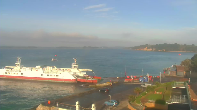 A ferry is docked at a busy harbor with water surrounding it. The ferry is white with red trim and has a visible control tower. In the foreground, there are vehicles parked along the waterfront, alongside a path with greenery and flower beds. In the background, small boats navigate the water, while a hillside with trees rises on the right side. The weather is clear with a blue sky and some clouds.