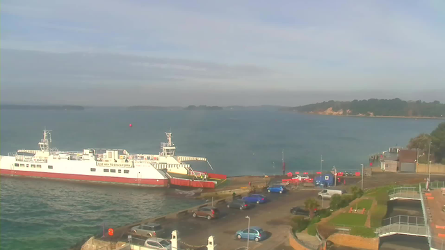 A ferry is docked at a pier beside a calm blue body of water. Several parked cars are visible in the foreground, with a few scattered people near the water's edge. In the background, a wooded shoreline rises gently, and the sky is light with some clouds. The ferry has a white and red color scheme, and there is a buoy in the water nearby.