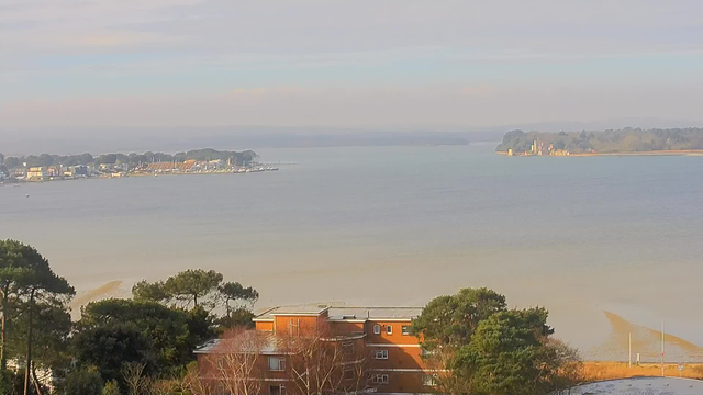 A calm body of water is visible, with a shoreline lined by trees and buildings. In the distance, a marina with boats is shown, along with a small island featuring structures on it. The sky is partially cloudy, with soft light illuminating the scene.