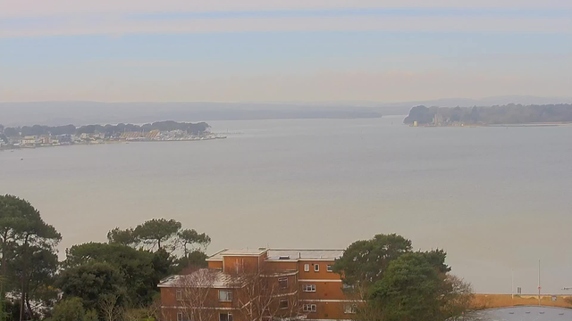 A serene view of a large, calm body of water, stretching across the image. In the foreground, there is a brown building surrounded by green trees. The water reflects soft, muted colors of blue and gray, with a faint haze in the distance. On the opposite shore, there are outlines of buildings and trees, with a marina visible where several boats are docked. The sky above is mostly clear with a gradient from pale blue near the horizon to soft white hues.