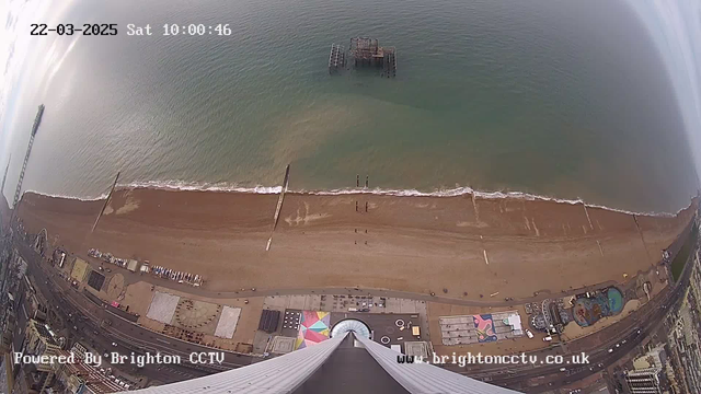 Aerial view of a beach with light brown sand and gentle waves lapping at the shore. In the foreground, there are several structures and a large circular area with a colorful design. To the left, a pier extends into the water, while a partially submerged structure is visible further out. The ocean appears calm and blue, and the scene is captured from a high vantage point, looking straight down. The date and time are displayed at the top of the image.