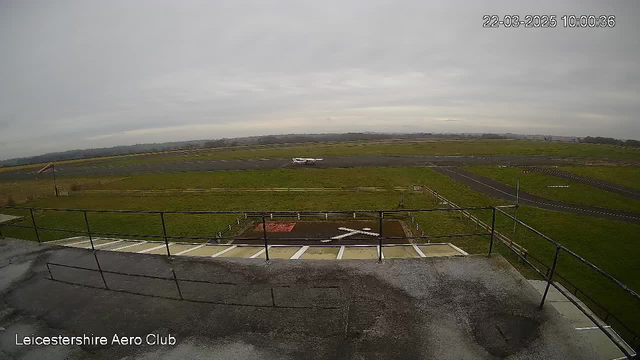 A vast, open landscape is captured from a high vantage point. The scene shows a grassy area leading to a runway at an aeroclub. In the distance, there are patches of low hills under a cloudy sky. A small white airplane is on the runway, which is marked with adjacent lines and a cross shape painted on the ground. A railing and some stairs are visible in the foreground, suggesting a viewing platform or observation area. The overall atmosphere is quiet and tranquil.