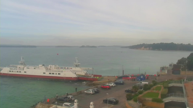 A view of a harbor with a white and red ferry docked at the edge of the calm water. The ferry is near a pathway leading to a small parking area filled with cars. On the left side, there are a few people standing by the waterfront, while in the background, the shoreline features green hills and distant trees under a light gray sky. The water is a mix of blue and green hues. In the foreground, there are landscaped areas with grass and decorative paths.