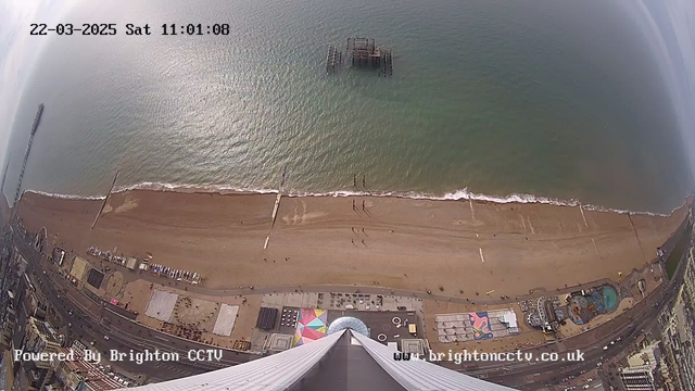 Aerial view of a beach on a clear day. The beach features golden sand with a few people walking along the shore. To the left, a long pier stretches out into the sea, and a sunken pier structure can be seen in the water. Various amusement park rides and attractions are located along the beachfront. There are colorful patterns on the ground, likely from beach facilities. The sky appears mostly clear, with some clouds.