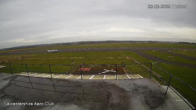 A view from a webcam overlooking an airfield on a cloudy day. In the foreground, there is a small elevated platform with a railing. To the right, a light aircraft is positioned on the runway, preparing to take off or land. There are a few people near a marked area on the ground, which features a large white "X." The landscape around the airfield is mostly grass with a few scattered structures in the distance under an overcast sky. The timestamp in the corner indicates the image was taken at 11:00:51 on March 22, 2025.
