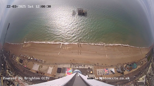 An aerial view of a sandy beach along the coastline, with gentle waves lapping at the shore. A pier extends into the sea, partially visible. The beach is dotted with a few people walking and sitting. Colorful beach structures and a carousel are seen on the left side of the image, while the ocean glimmers under sunlight. The sky is mostly clear with some wispy clouds. The bottom of the image shows part of a tall structure from which the photo is taken, tapering off towards the top.