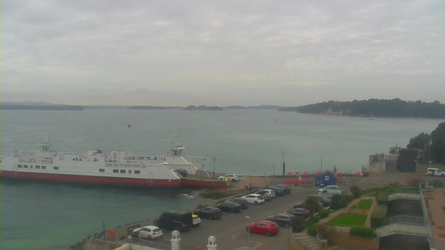 A ferry is docked at a harbor, with its white and red hull visible. The water reflects a grey sky filled with clouds, and distant islands are visible on the horizon. A variety of cars are parked along the waterfront, and some people can be seen near the ferry. In the foreground, there is greenery and landscaping near the parking area, while a small blue building and construction barriers are also visible near the harbor.