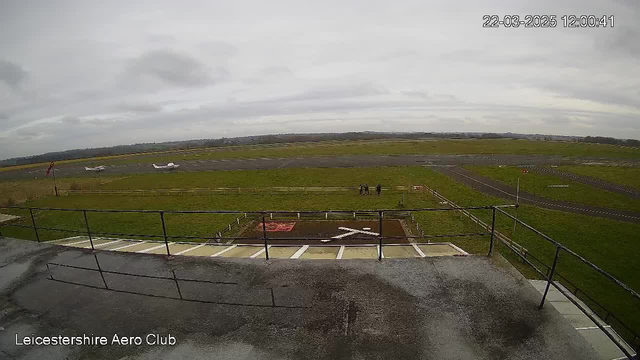 A view from a webcam at the Leicestershire Aero Club shows a grassy airfield under a cloudy sky. Two small aircraft are positioned near the runway, with one on the left and another farther down the runway. A group of three people stands on the grass near a marked section, which includes an "X" shape. A fence borders the airfield, and there are some signs visible in the background.