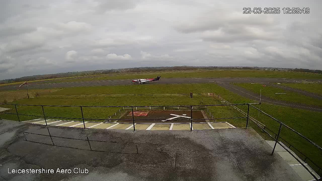 A cloudy sky is visible above a green field with a runway. A small aircraft, predominantly white with red and black accents, is positioned on the runway. In the foreground, there is a viewing platform with a railing, and a person stands on the ground near the edge of the grass. The area is relatively open with a few scattered objects, such as a windsock and markings on the ground.