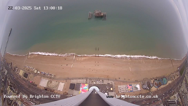 Aerial view of a beach and seafront, with golden sand stretching across the lower half of the image. Gentle waves are lapping at the shore, creating a frothy edge. In the distance, an old wooden pier juts out into the water. The area adjacent to the beach shows various structures and attractions, including colorful tents and buildings. The upper portion of the image presents a clear sky, blending into the calm blue-green sea. A timestamp in the top left corner indicates the date and time.