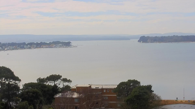 A serene view of a wide body of water with a calm surface, reflecting the sky. In the foreground, there are trees and the rooftops of buildings, predominantly brown and light-colored. To the left, there's a marina with several boats and structures, while the right side features a wooded area along the water's edge. The distant shoreline is lined with trees and hills under a light, partly cloudy sky.